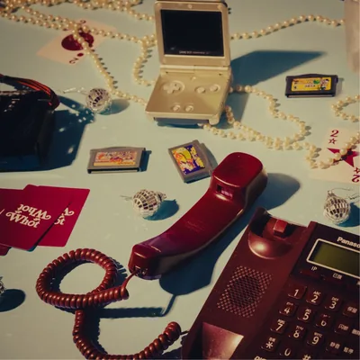Cover art: a red telephone handset, a Game Boy Advance SP, pearls and playing cards on a pale blue table