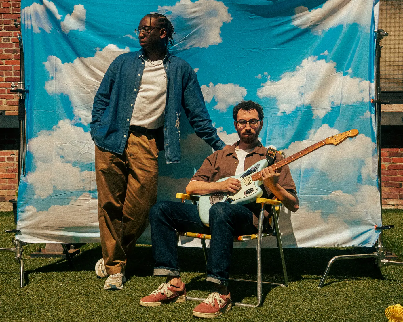 Two Post Modern Connection members, one standing and one seated with an electric guitar, in front of a cloud-print backdrop