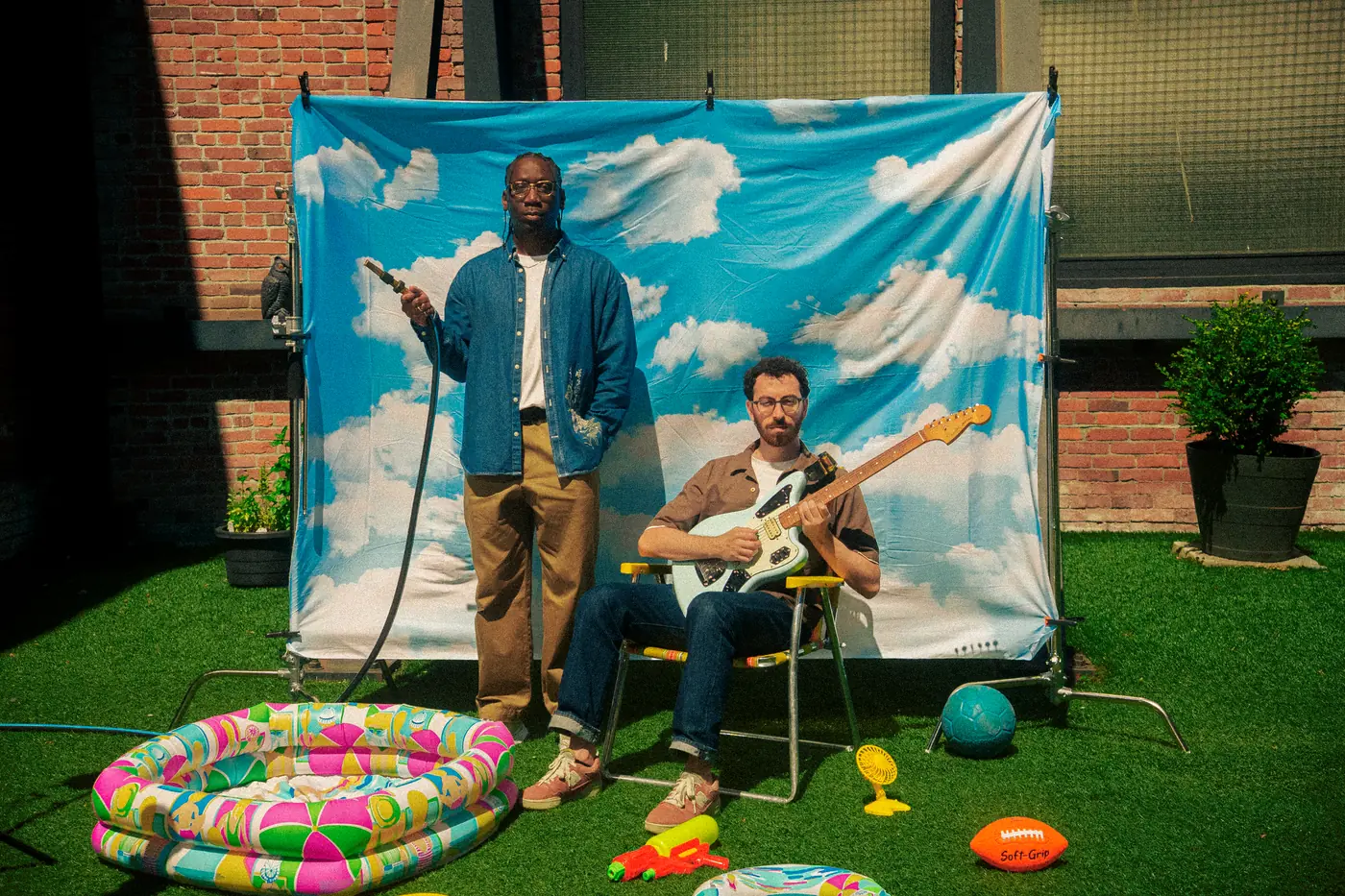 The two members of Post Modern Connection, one standing with a garden hose and one seated with an electric guitar, in front of a cloud-print backdrop in a Vancouver backyard